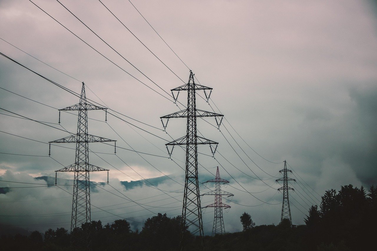 High-voltage transmission towers and power lines crossing open country at dusk, representing the grid cost of the AI data center build-out, October 2026